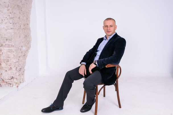 A man in a blue shirt, black trousers and a blue jacket sits on a black stool on a white background - Photo, Image