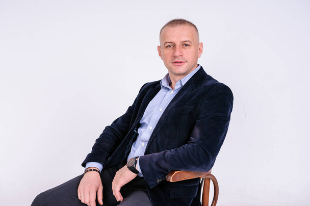 A man in a blue shirt, black trousers and a blue jacket sits on a black stool on a white background - Fotografie, Obrázek