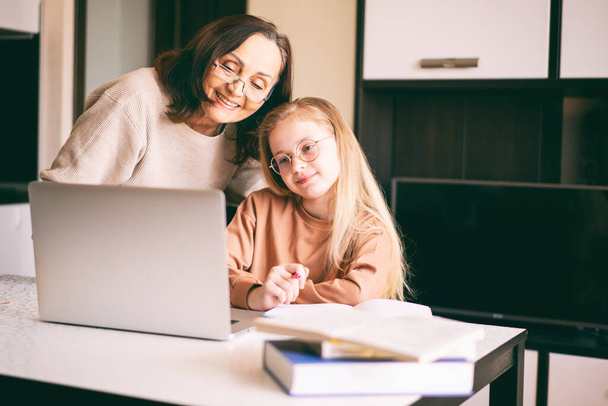 grandmother in eyeglasses helping her granddaughter with her homework. Beautiful 10-year-old girl doing homework on a laptop with the support of her grandma.. - Foto, Imagem