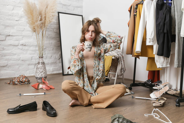 Stressed young woman sitting in the messy room full of clothes. Nothing to wear concept. - Photo, Image