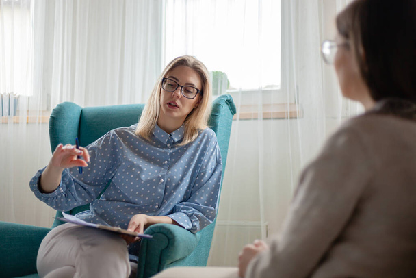 Woman psychotherapist consults patient sitting in armchair at home, writes on a clipboard - Fotografie, Obrázek