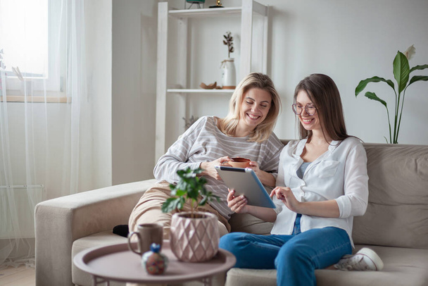 Two young women friends using tablet together, sitting on the couch, smiling and fun - Photo, Image