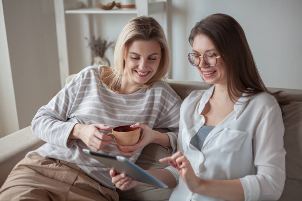Дві молоді жінки друзі використовують планшет разом, сидячи на дивані, посміхаючись і весело
 - Фото, зображення