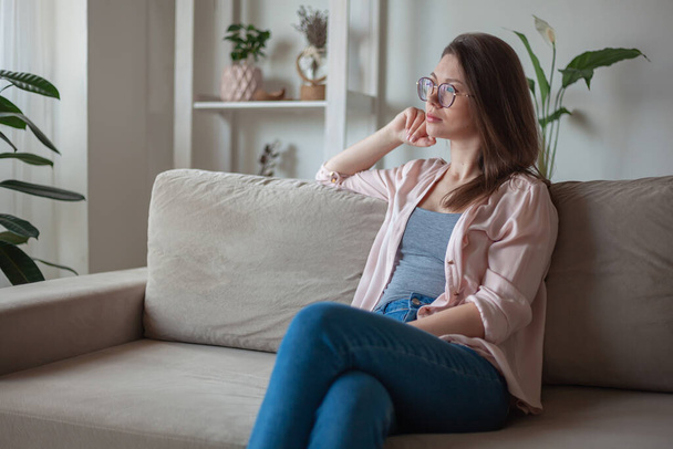 Young woman with eyeglasses sitting on sofa and looking the window thoughtfully - 写真・画像