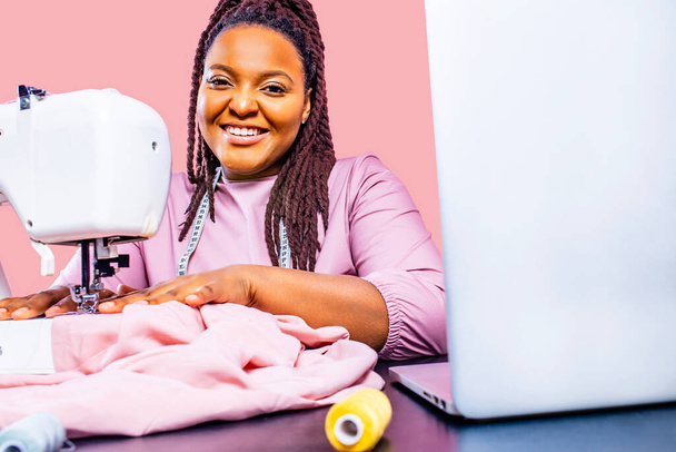 african tailor with afro dreadlocks pigtails sews clothes on sewing machine at tailor office pink wall background - Фото, изображение
