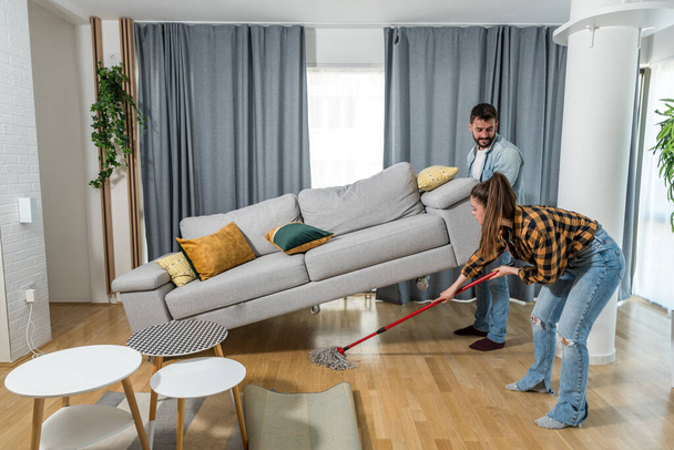 Young couple in their apartment a woman wipes and cleaning the floor and a man helping her by rising up the sofa bed with his hands so that she can clean the floor from dust and dirt - 写真・画像
