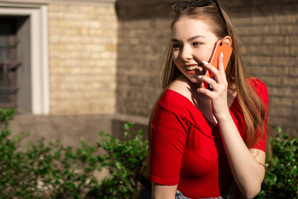 Street candid portrait of a young woman in red talking on the phone and smiling. Hot ummer day with hard sun - Photo, Image