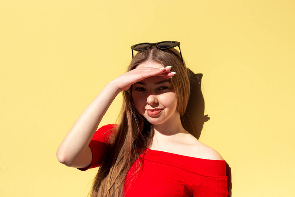 Retrato candid da rua de uma mulher nova no vermelho que está na frente da parede amarela e que esconde sua cara do sol. Luz solar dura, sombras duras - Foto, Imagem