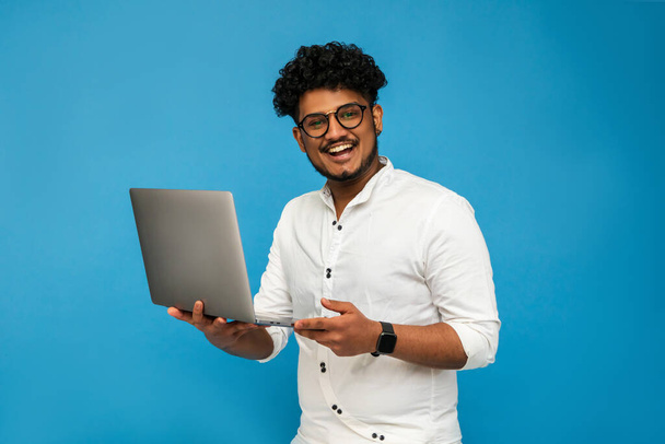Indian manager, Indian freelancer isolated on blue background, studio photo, holding laptop and hands and smiling looking at camera. Invitation to work - Fotó, kép