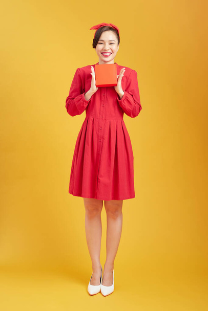 Красива весела жінка в червоній сукні дарує вам червоний подарунок на жовтому тлі
 - Фото, зображення