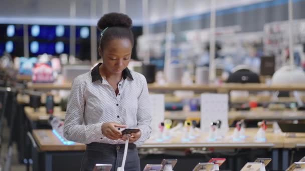 mladá afro-americká žena vybírá mobilní telefon, prohlížení exponátů v obchodě s digitálním vybavením, dáma kupuje smartphone v maloobchodě - Záběry, video