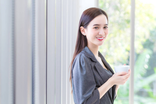 Aasian työskentelevät nainen pukeutuu harmaa mekko seisoo ja pitää kahvikupin käsissään aamulla toimistossa. - Valokuva, kuva