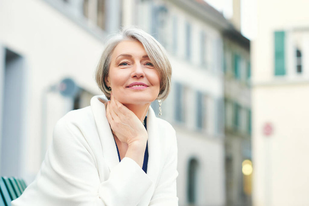 Portrait extérieur de belle et élégante femme de 55 à 60 ans, vêtue d'une veste blanche, assise sur un banc à l'extérieur - Photo, image
