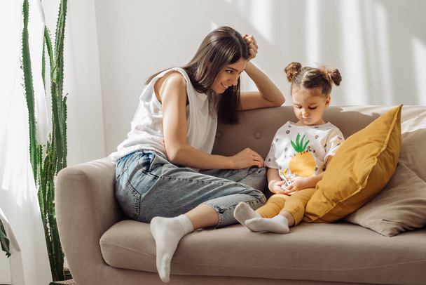A dark-haired young mixed-race woman plays on the couch with her little daughter.Home interior design with houseplants.Time together.Family concept and Mother's Day concept. - Photo, image
