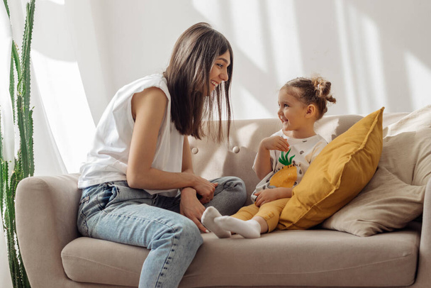 A dark-haired young mixed-race woman plays on the couch with her little daughter.Home interior design with houseplants.Time together.Family concept and Mother's Day concept. - Photo, image