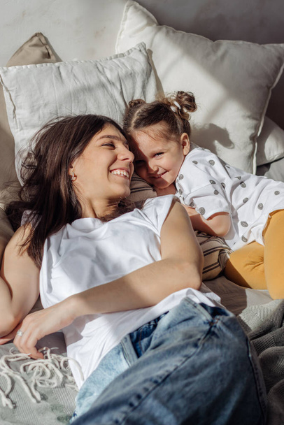 Tummahiuksinen nuori sekarotuinen nainen makaa sängyssä pienen tyttärensä kanssa.Sisustussuunnittelu.. - Valokuva, kuva