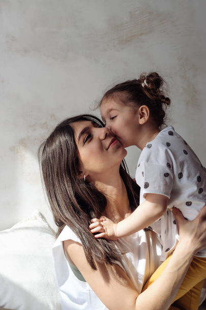 A dark-haired young mixed-race woman playing on the bed with her little daughter.The girl kissing her mother.Home interior design.Time together.Family concept and Mother's Day concept. - Foto, immagini