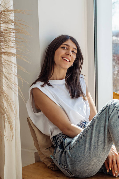 A dark-haired mixed-race girl sitting on the windowsill and enjoying the sunny day.Home interior design.View of the city outside the window.The concept of relaxation and good mood. - Valokuva, kuva