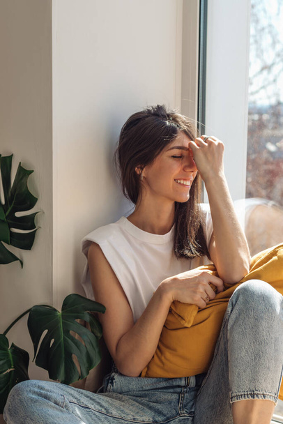 A dark-haired mixed-race girl sitting on the windowsill and enjoying the sunny day.Home interior design.View of the city outside the window.The concept of relaxation and good mood. - Foto, imagen