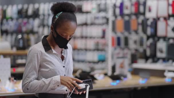 Αφρο-αμερικανική γυναίκα με μάσκα προσώπου επιλέγει σύγχρονο smartphone στο κατάστημα ψηφιακού εξοπλισμού, προβολή νέων μοντέλων στην αίθουσα συναλλαγών πριν από την αγορά - Πλάνα, βίντεο