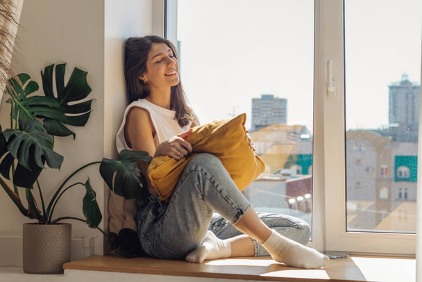 A dark-haired mixed-race girl sitting on the windowsill and enjoying the sunny day.Home interior design.View of the city outside the window.The concept of relaxation and good mood. - Zdjęcie, obraz