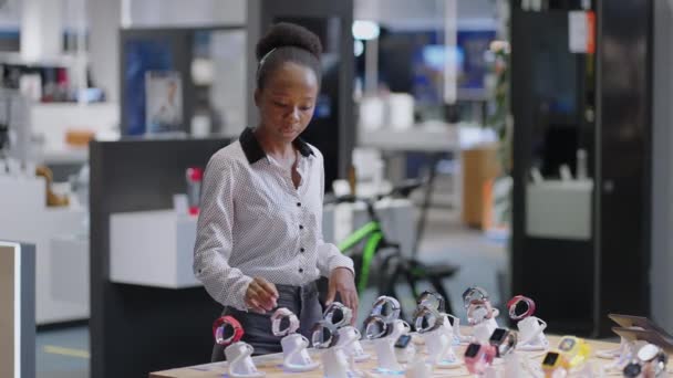 молодая афро-американская покупательница выбирает умные часы в магазине цифрового оборудования, просматривая модели на демонстрационной витрине - Кадры, видео