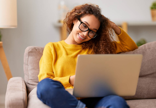 Young beautiful african american woman wearing eyeglasses working remotely on laptop while sitting on sofa at home, cheerful female student studying online. Distance education and freelance concept - Fotoğraf, Görsel