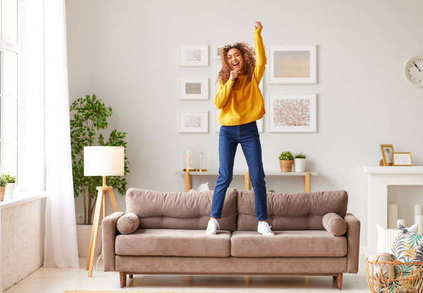 Pretending to be famous singer. Overjoyed afro american teenage girl in casual wear singing and dancing on sofa in modern living room at home. Young happy female enjoying carefree and leisure weekend - Foto, imagen