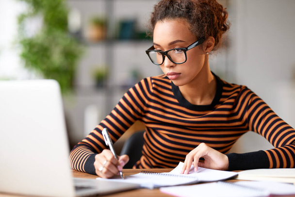 Mujer afro-americana enfocada haciendo algunas notas y mirando la pantalla del portátil mientras está sentada en su lugar de trabajo y trabajando remotamente. Estudiante de raza mixta concentrada que estudia en línea en casa - Foto, Imagen