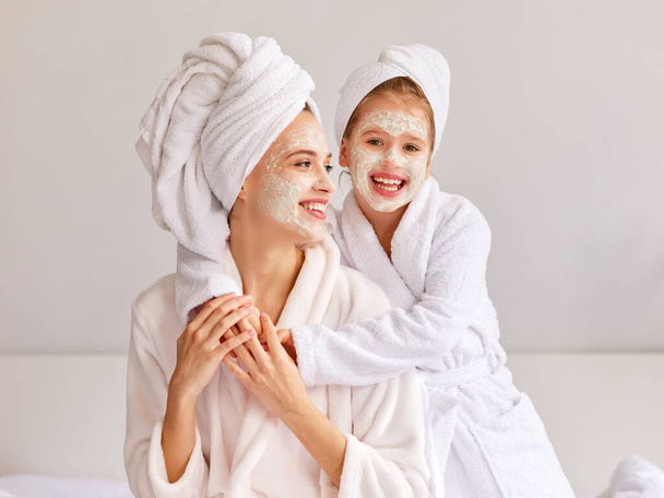 Cheerful girl in bathrobe and towel smiling and embracing young woman with moisturizing mask while resting on bed at home - Photo, Image