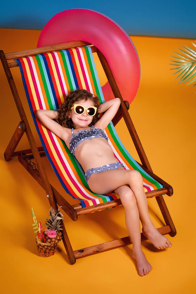 Smiling girl in lilac swimsuit and sunglasses lying in rainbow deck chair with crossed legs and sunbathing - Foto, Bild