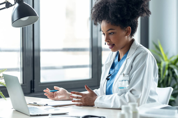 Shot of attractive afro female doctor talking while explaining medical treatment to patient through a video call with laptop in the consultation. - 写真・画像