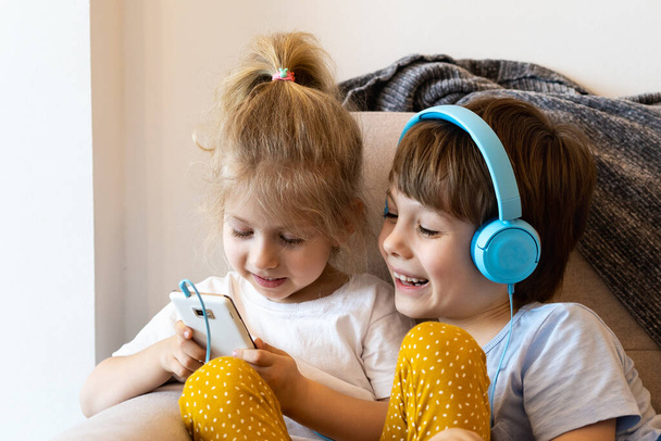 Hermano y hermana se sientan en casa en el sofá con teléfonos inteligentes, chica en auriculares, concepto de niños y la tecnología moderna. - Foto, imagen