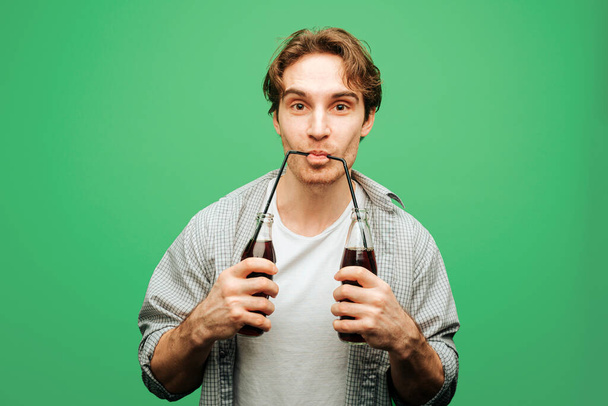 Man holds glass bottles and drinks from two straws, isolated on green background - Foto, Imagen