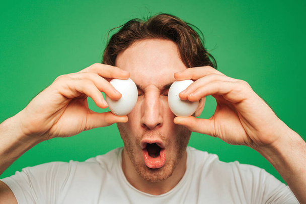 Young man covers eyes up by eggs, isolated on green background - Photo, Image