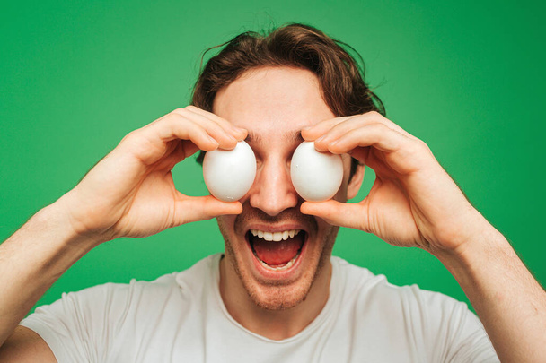 若い男は緑の背景に隔離された卵で目を覆う - 写真・画像