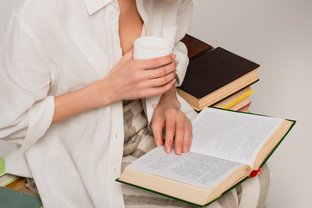 cropped view of woman reading book and holding cup of coffee isolated on grey  - Photo, Image