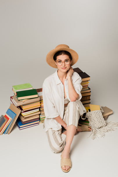 pełna długość zadowolonej kobiety w kapeluszu i okularach siedzi w pobliżu stosu książek na białym - Zdjęcie, obraz