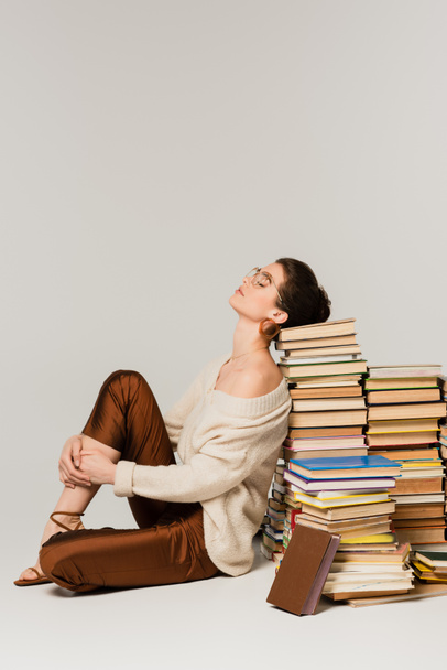 Gözlüklü ve süveterli genç bir kadın beyaz kitaplara yaslanıyor. - Fotoğraf, Görsel