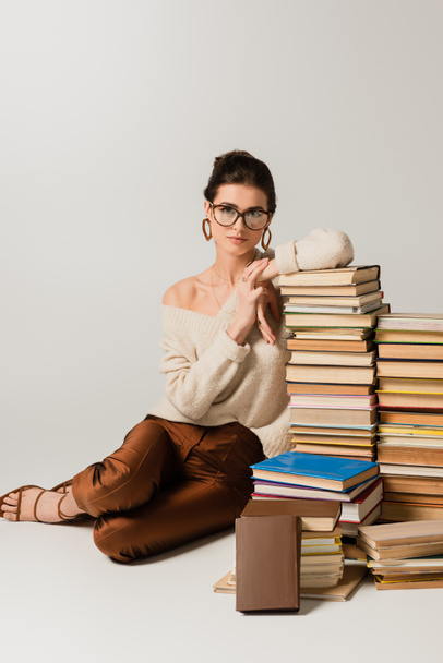 full length of young woman in glasses and earrings leaning on stack of books on white - Zdjęcie, obraz