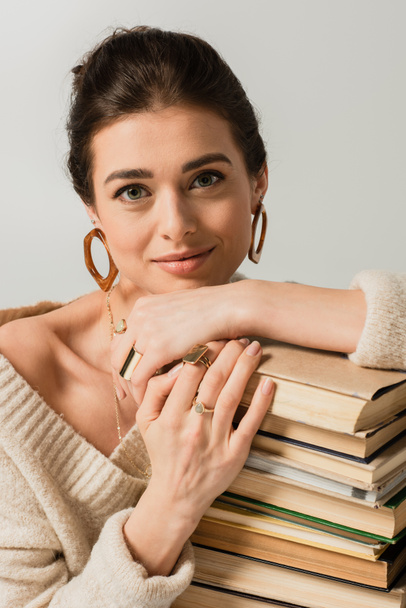 smiling young woman in earrings leaning on stack of books isolated on white - Photo, image