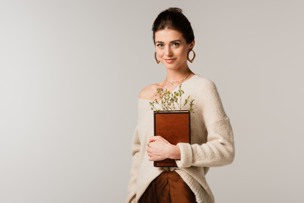 pleased young woman holding book with wildflowers isolated on grey - Photo, Image