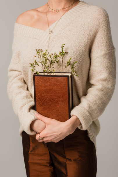 cropped view of young woman holding book with wildflowers isolated on grey - Fotó, kép