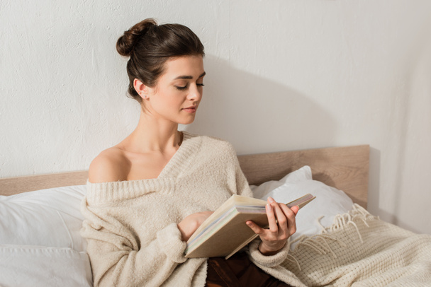 brunette jeune femme lecture livre tout en se reposant sur le lit à la maison - Photo, image
