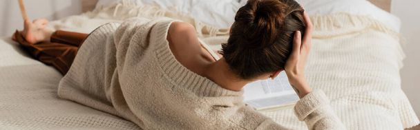 back view of young woman reading book while resting on bed at home, banner - Фото, изображение
