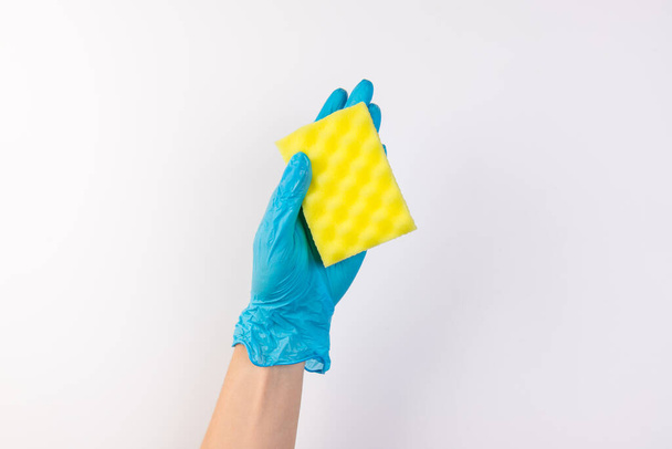 Main féminine en gants bleus pour le nettoyage et les produits de nettoyage pour la maison tient une éponge jaune sur un fond blanc isolé - Photo, image