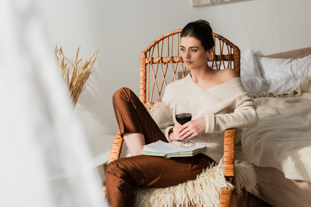 pretty young woman sitting in wicker rocking chair with book and glass of wine - Photo, Image