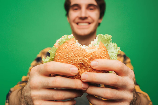 Close up young man holds burger with pleasure, isolated on green background - Photo, Image