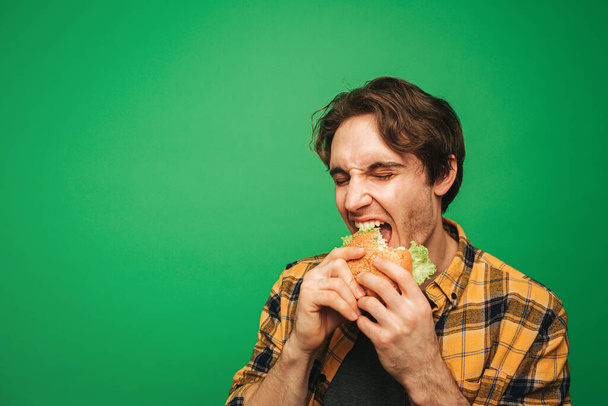 Giovane uomo mangia hamburger con piacere, isolato su sfondo verde - Foto, immagini