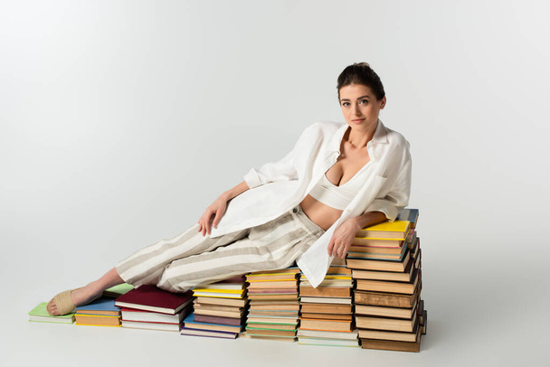 πλήρες μήκος της μελαχρινής νεαρής γυναίκας με σανδάλια ξαπλωμένη σε σωρό από βιβλία σε λευκό  - Φωτογραφία, εικόνα